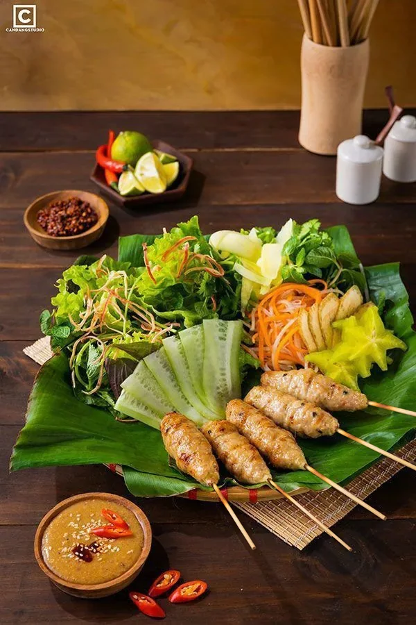 Mẹt nem nướng xiên que tre bày trên lá chuối cùng rau sống, khế, dưa leo và chén nước chấm