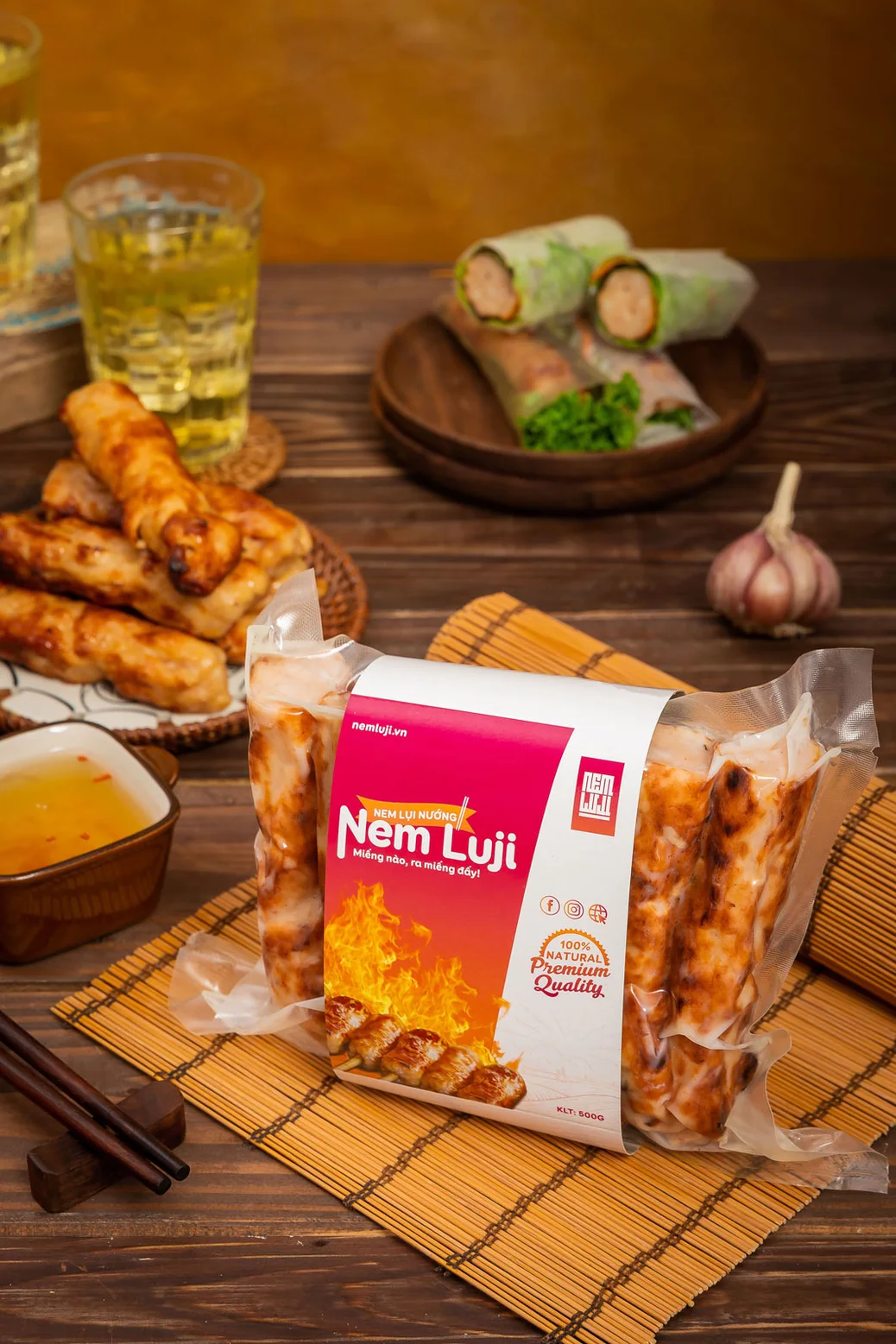 Gói nem nướng Nem Luji 500g trên mành tre, cạnh đĩa nem đã nướng và chén nước chấm
