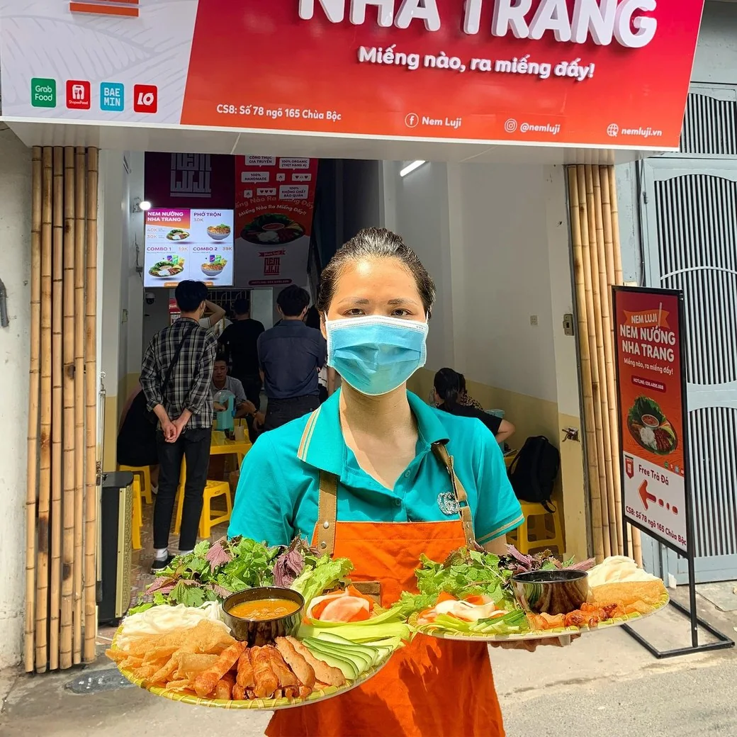 Nhân viên áo xanh bưng hai mâm nem nướng trước cửa quán
